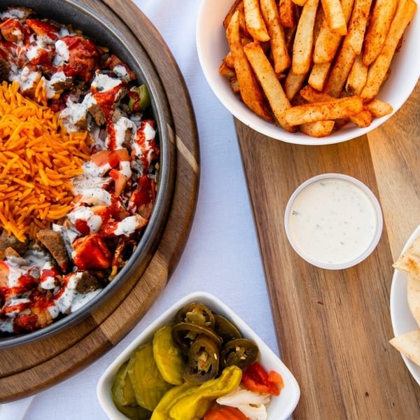 A vibrant platter of chicken and lamb over rice with sauces, surrounded by fries, chickpeas, pita, and pickles.