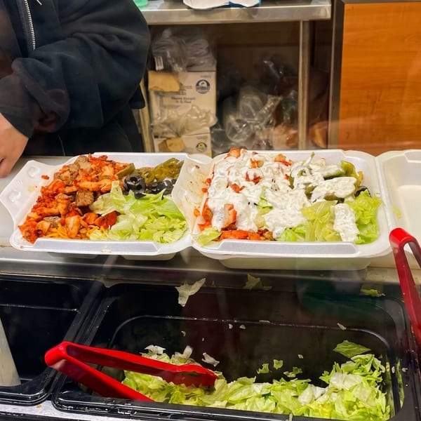 Two styrofoam containers filled with chicken, lettuce, olives, and sauce at a food counter.