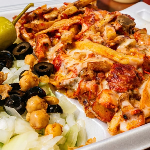 A white styrofoam container holds cheesy fries with chicken, a side salad, chickpeas, olives, and a pepper.