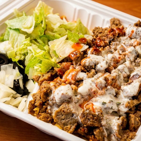 Takeout tray filled with seasoned meat, creamy sauce, lettuce, onions, and black olives on a wooden table.