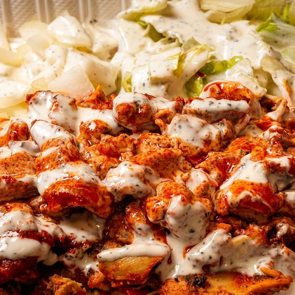 Grilled chicken pieces drizzled with spicy red and creamy white sauces over yellow rice, lettuce, and onions in a takeout tray.