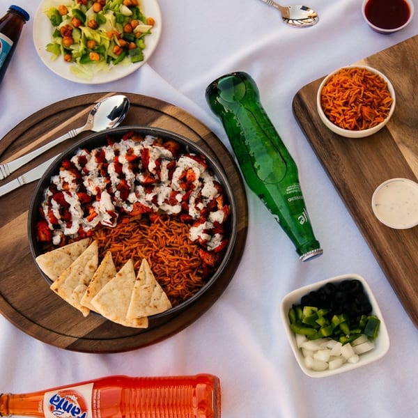 Overhead view of a chicken and rice platter with pita, salads, sauces, and soda bottles on a white tablecloth.