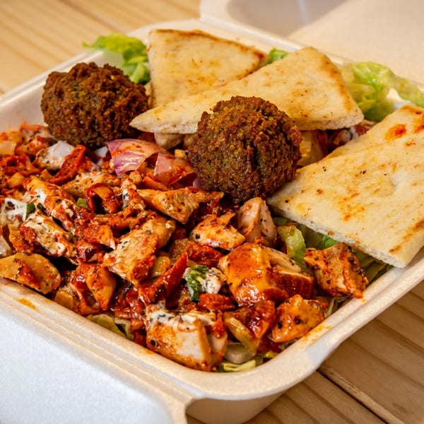 Takeout container filled with spicy grilled chicken, falafel, pita wedges, salad, and creamy sauce on a wooden table.