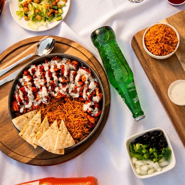 Overhead restaurant table spread featuring saucy grilled meat, spiced rice, flatbread, salads, pickles, vegetables, and bottled drinks.