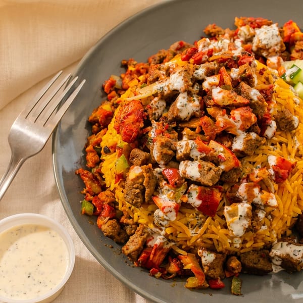 Mediterranean-style platter with yellow rice, seasoned meat, roasted vegetables, salad, creamy sauce, and fork.
