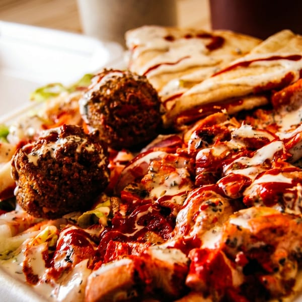 A close-up of a takeout container filled with falafel, chicken, pita bread, and sauces.